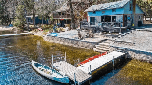 A serene lakeside view featuring a dock, kayaks, and a cozy blue house surrounded by trees.